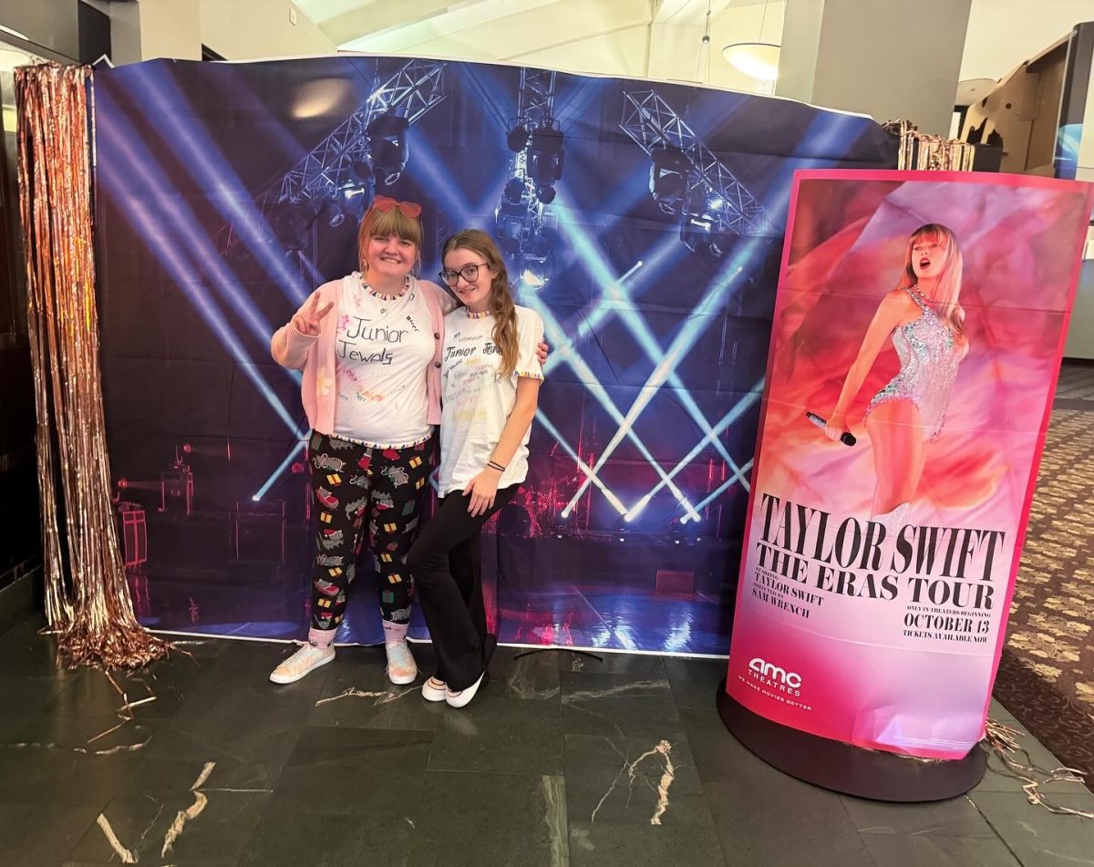Swift poses with cousin Grace Kelly in matching “Junior Jewels” shirts in front of Eras Tour movie poster
